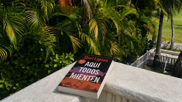 Libro Aquí todos mienten, de Shari Lapena, sobre una balaustrada blanca con palmeras tropicales al fondo.