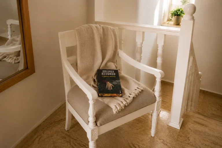 Libro El guardián invisible de Dolores Redondo apoyado sobre una silla blanca con manta beige en un rincón luminoso junto a una barandilla.