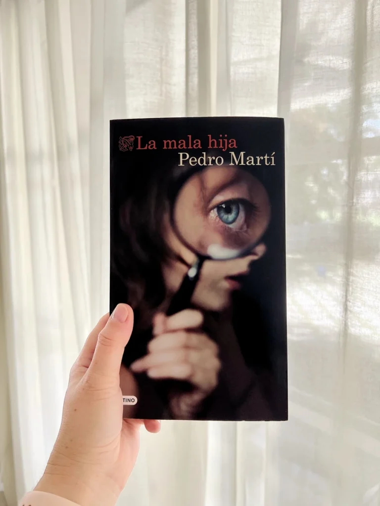 Portada del libro La mala hija, de Pedro Martí, sostenido con una mano delante de unas cortinas blancas iluminadas por luz natural.
