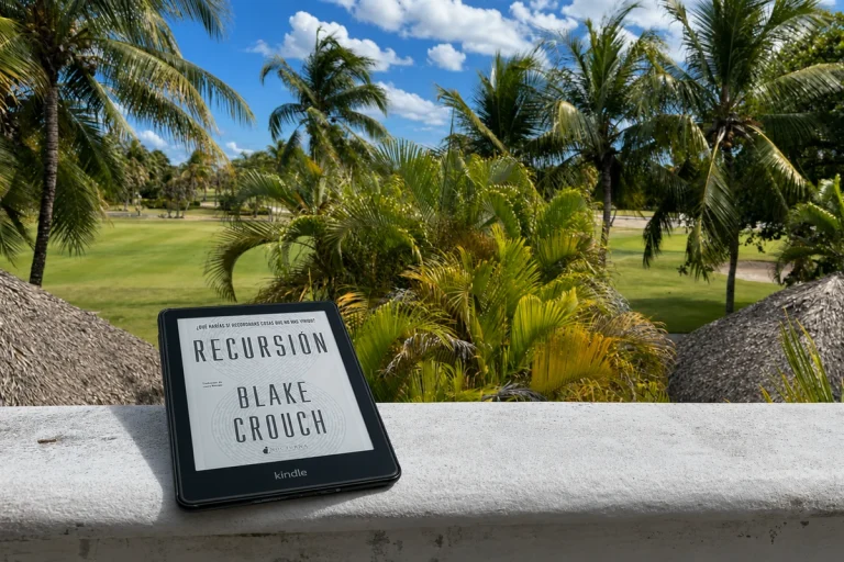 Kindle con Recursión de Blake Crouch sobre una balaustrada blanca con paisaje tropical al fondo.